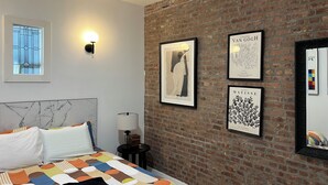 A hotel room with a brick wall, a bed with a colorful quilt, a nightstand with a lamp, and artwork including pieces by Vincent Van Gogh and Henri Matisse.