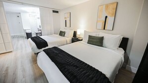 Habitación de hotel con dos camas, un escritorio y una silla. Se observan obras de arte en las paredes y una ventana que deja entrar luz natural.