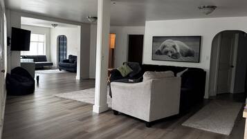 En rummelig opholdsstue med en grå sofa, et fladskærms-tv monteret på væggen og et stort vindue, der lukker naturligt lys ind.