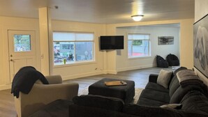 Un salon comprenant un grand canapé, une table basse et un téléviseur à écran plat fixé au mur. De grandes fenêtres laissent entrer la lumière naturelle et offrent une vue sur l’extérieur.