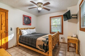 Ein Schlafzimmer mit einem hölzernen Bett, einem Deckenventilator, einem an der Wand befestigten Fernseher und einem Fenster mit Jalousien.