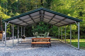 Ein überdachter Picknickbereich mit Tisch und Stühlen, umgeben von Kies und Bäumen.