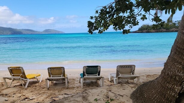 A beach with four sun loungers under a tree, a clear blue sea, and distant mountains. - Villa between sea and mountain with beautiful places, live the adventure and romance. (Monte Rojo)