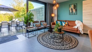 Un salon moderne avec un canapé, une table basse et des fauteuils. Une grande fenêtre offre une vue sur les arbres, et une terrasse aménagée comprend une table et des chaises.