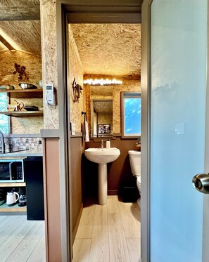 Eine kleine Küche mit Mikrowelle, Kaffeemaschine und Regalen. Durch eine geöffnete Tür ist ein Badezimmer mit Waschbecken, Toilette und Spiegel zu sehen.