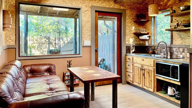 A cozy room with a leather sofa, a wooden table, a kitchen with wooden cabinets, and a large window offering a view of trees. - Pet Friendly Fully Fenced Cozy Casita near Sedona! (Lake Montezuma)