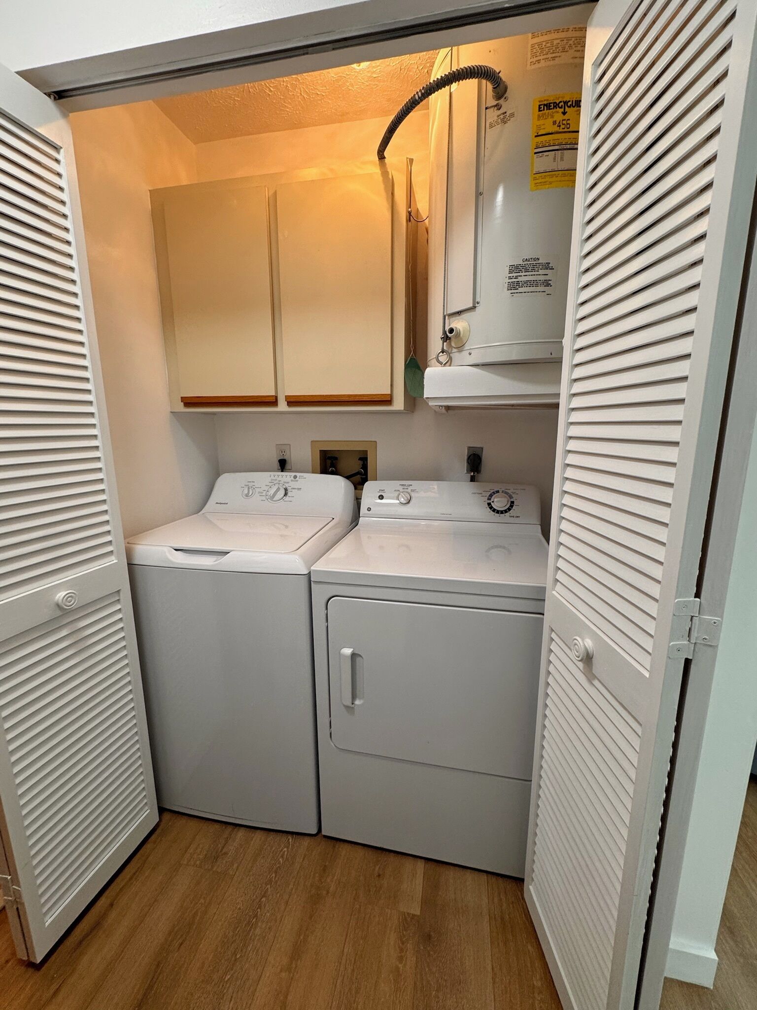 A compact laundry room with two front-loading washers and dryers, wooden cabinets above, and a caution sign on the wall.