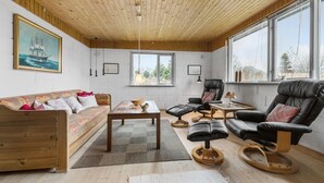 Un salon chaleureux avec un plafond et des murs en bois, un canapé, une table basse et un fauteuil en cuir. Deux fenêtres laissent entrer la lumière naturelle et offrent une vue sur l’extérieur.