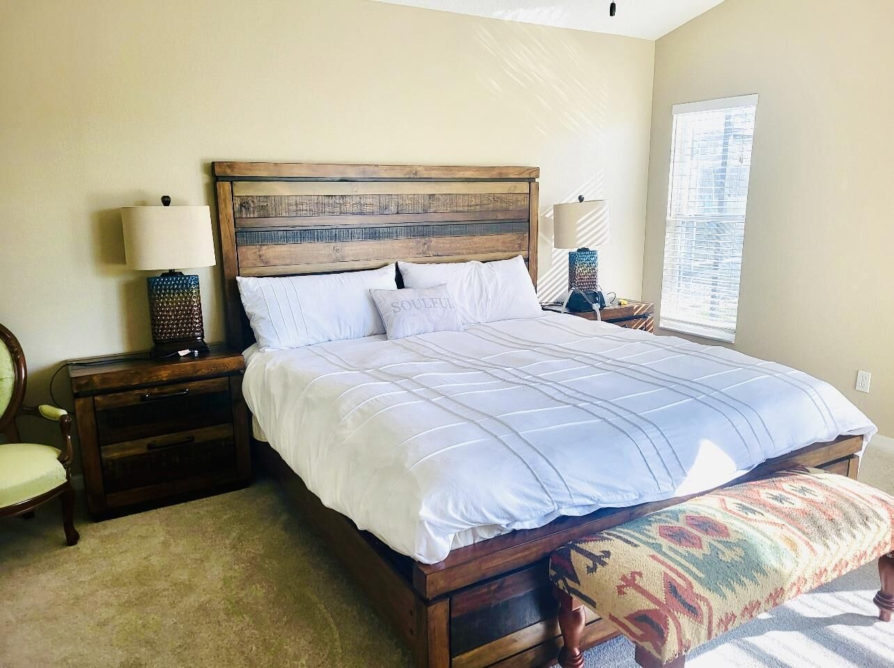 A bedroom with a wooden headboard, a large bed with white bedding, a bedside table with a lamp, and a window with blinds.
