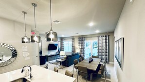 A modern kitchen with a central island, a dining area with a table and chairs, a blue sofa, and a TV mounted on the wall. - Beautiful Condo at Reunion (Kissimmee)