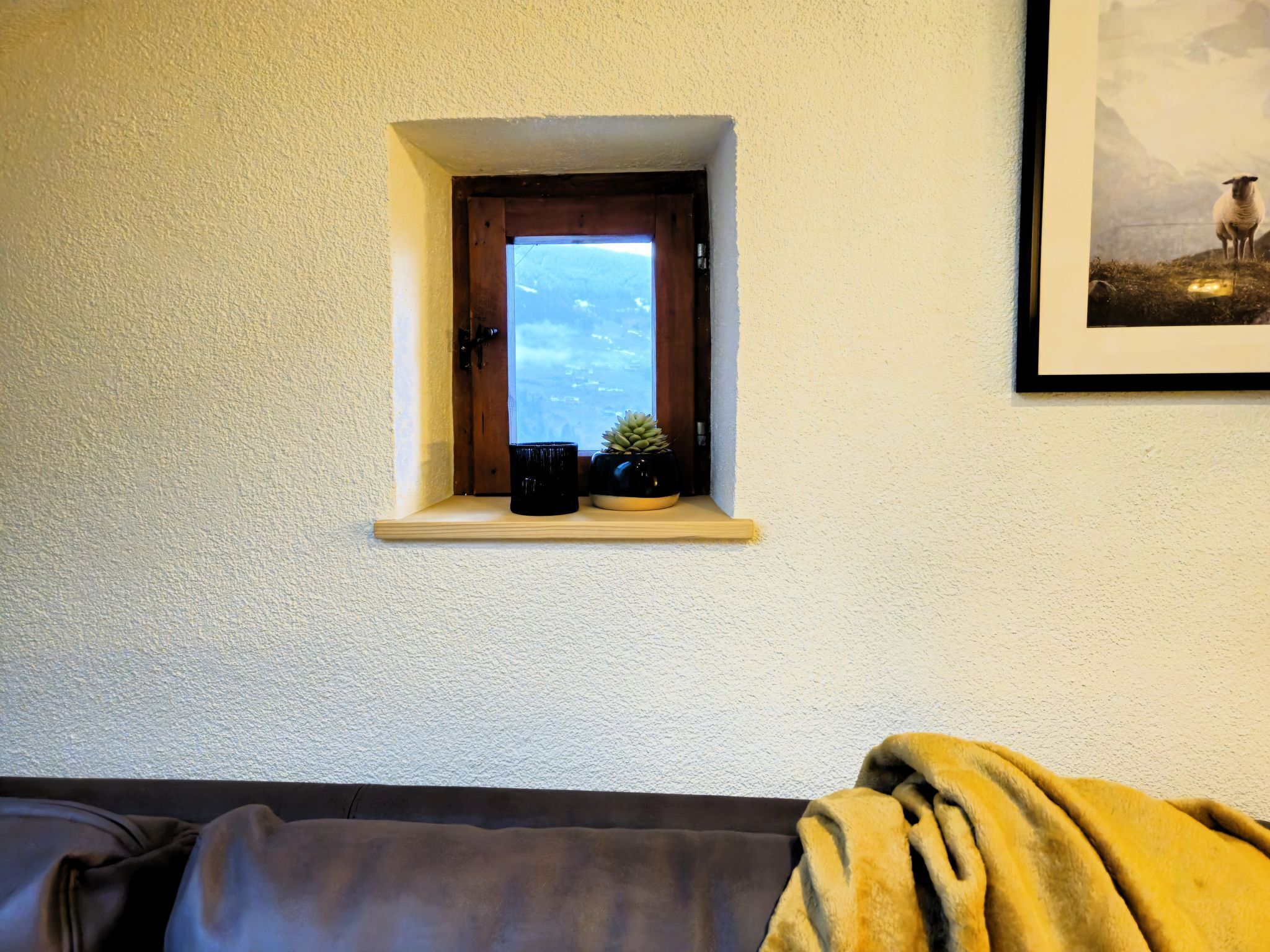 Una piccola finestra con vista su un paesaggio montano, uno scaffale con due piante in vaso e un quadro appeso al muro.