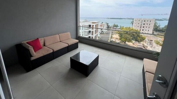A balcony with a beige sofa and a black coffee table, overlooking a cityscape and water. - 2-bedrooms condo with spectacular Ocean View, WiFi, AC in delightful Montego Bay (Montego Bay)