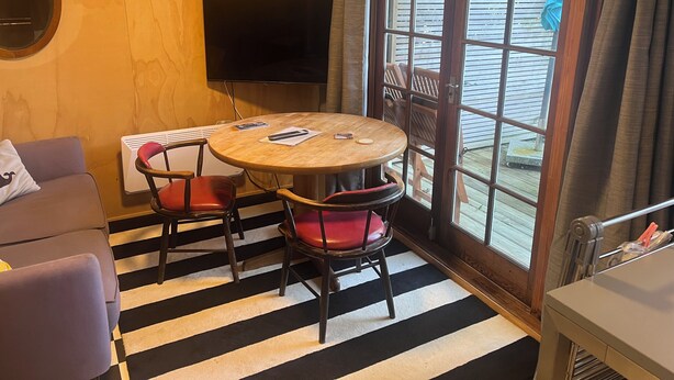Ein Zimmer mit einem runden Holztisch, zwei roten Stühlen, einem an der Wand befestigten Fernseher und einem großen Fenster mit Blick ins Freie.