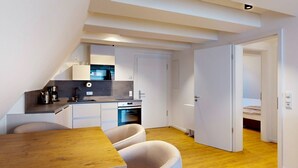En kompakt køkken med moderne hårde hvidevarer, et spiseområde med et træbord og stole, og et soveværelse, der kan anes gennem en åben dør.