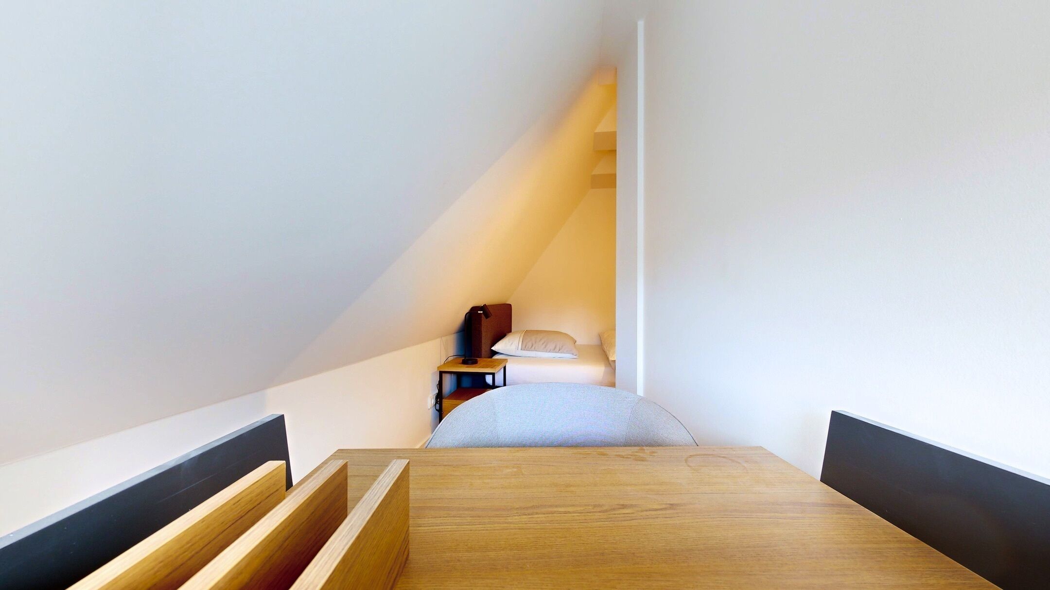 Una habitación estrecha y con techo inclinado, con una cama de madera al final, una mesita de noche pequeña y una silla.
