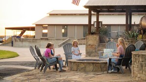 An outdoor patio area with stone seating and a fire pit, surrounded by a building with a metal roof and an American flag. - Cozy studio with queen pullout sofa on scenic working ranch in Fort Worth. (Fort Worth)
