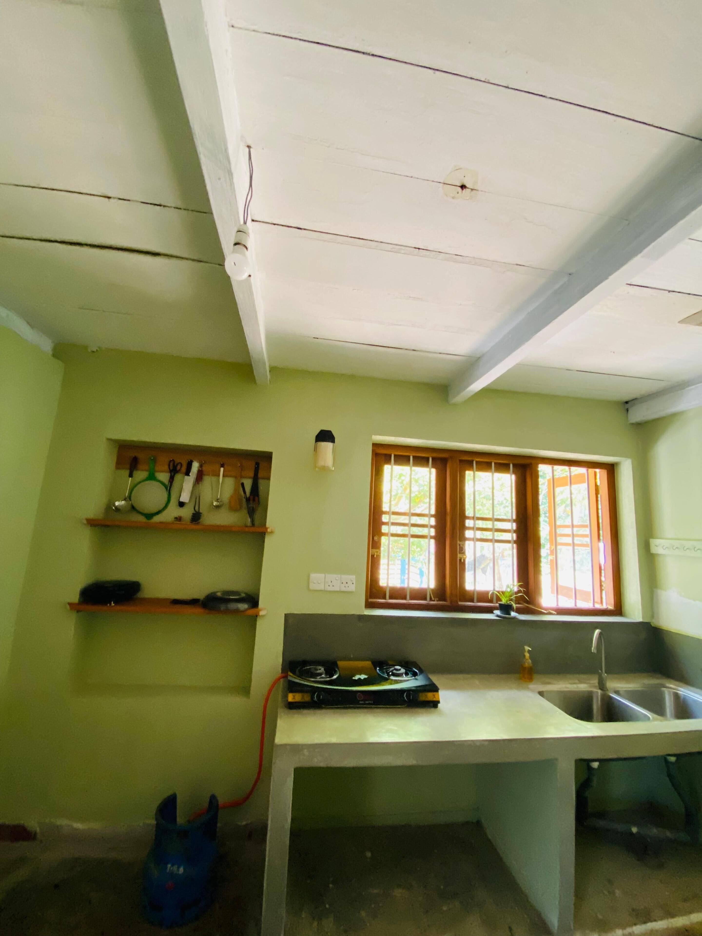 Una cocina con estufa a gas, lavamanos y ventana con rejas. Hay estantes con utensilios de cocina y una planta en maceta en el alféizar.