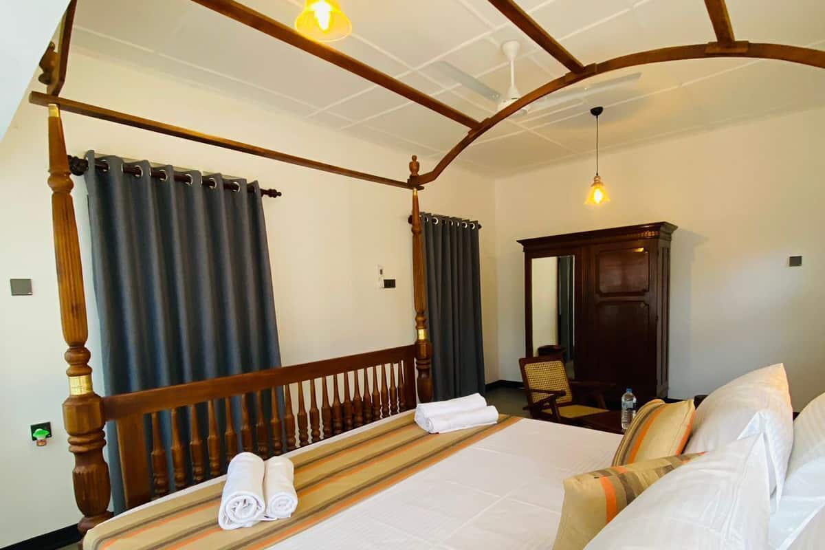 A four-poster bed with a wooden frame, white linens, and a striped bed runner. There is a wooden nightstand with a mirror and a lamp. The room has white walls and a ceiling fan.