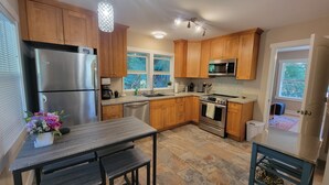 A modern kitchen with wooden cabinets, stainless steel appliances, a dining area with a bench, and large windows providing natural light. - Charming Lake Cottage+Private Dock+AC—Bring your paddles floats & fishing rods! (Sedro-Woolley)