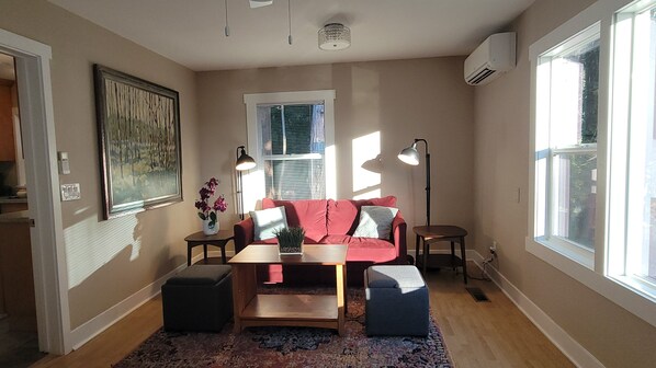 Ein gemütliches Wohnzimmer mit einem roten Sofa, einem Couchtisch, einem kleinen Beistelltisch mit Lampe und einem Fenster mit Jalousien.