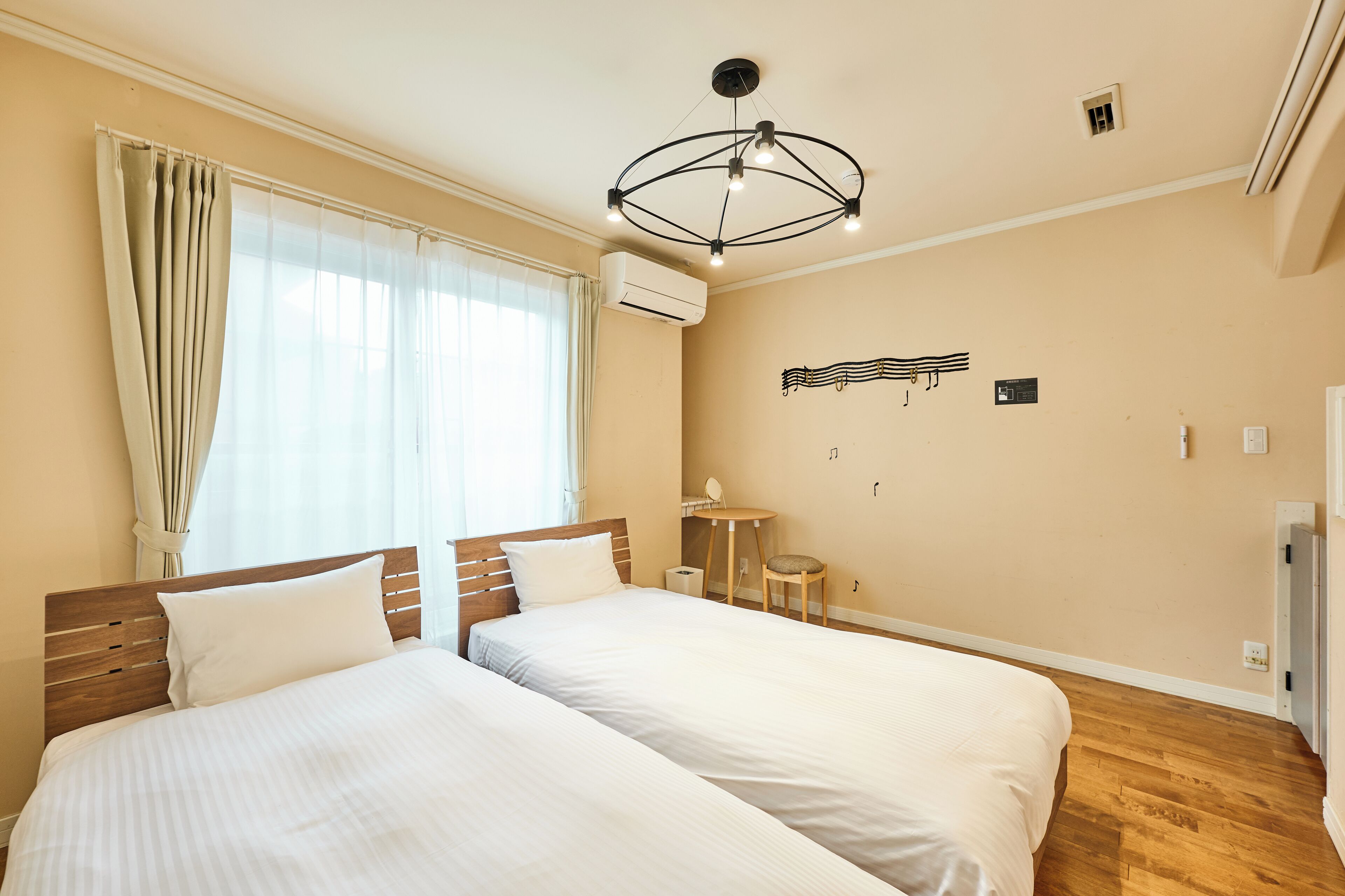 Habitación de hotel con dos camas, una lámpara en el techo, un equipo de aire acondicionado, una mesita con una silla y un atril sujeto a la pared.