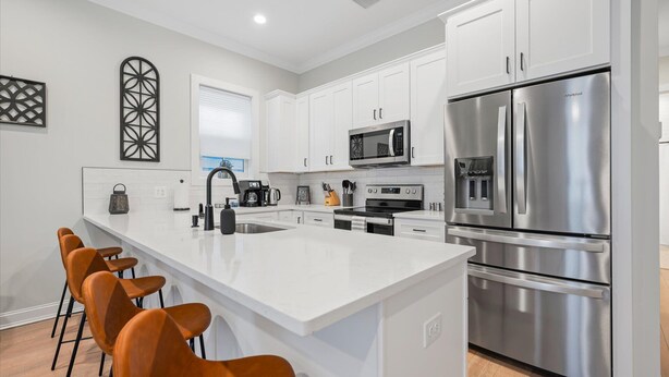 Una cocina moderna con muebles blancos, electrodomésticos de acero inoxidable, una isla central con taburetes y un microondas sobre la estufa.