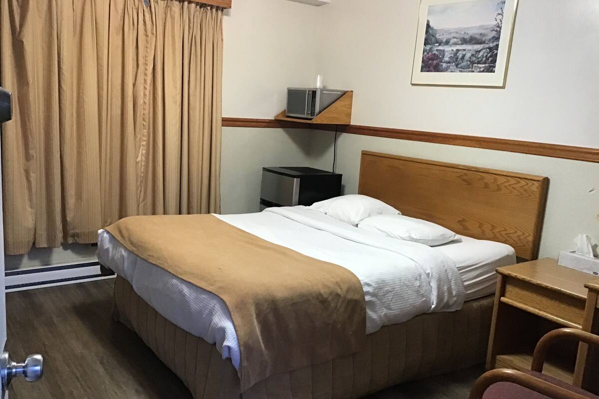 A hotel room with a bed, a nightstand, a chair, a picture on the wall, and a window with curtains.