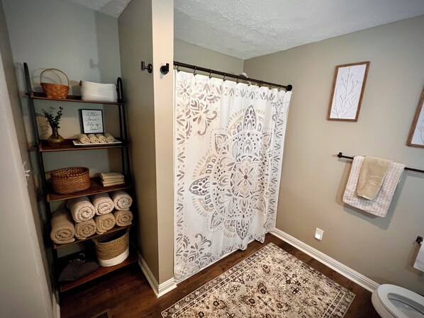 Une salle de bain avec un rideau de douche, des toilettes, un tapis et des étagères où sont rangées des serviettes et des paniers.