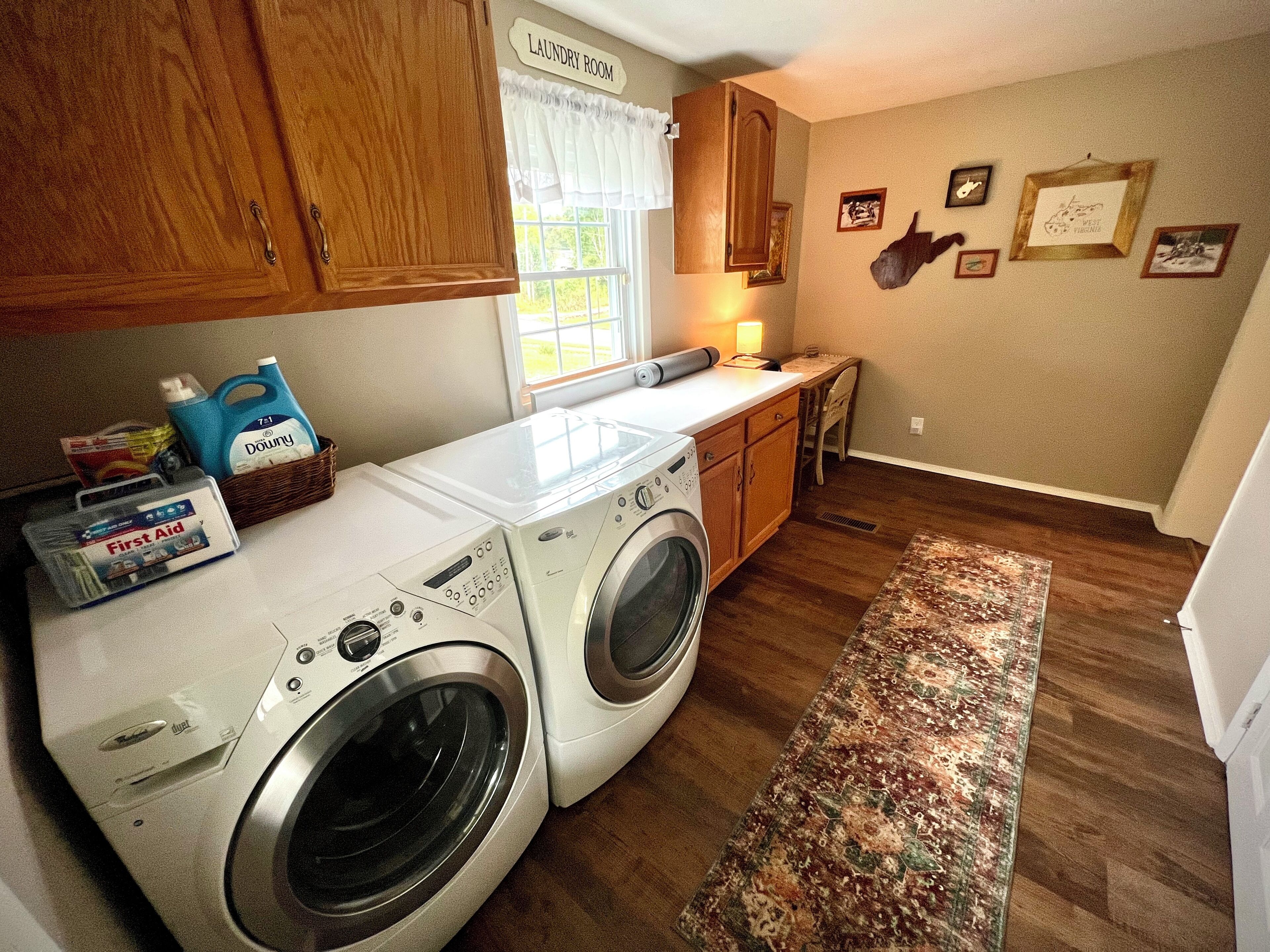 A laundry room with two front-loading washers, wooden cabinets, a window with white curtains, a small table with a lamp, and a rug on the floor.