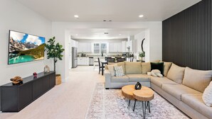 Una sala de estar moderna con un sofá, una mesa de centro y un televisor grande. Se observa una cocina al fondo con armarios y un comedor.