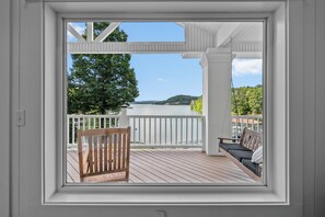 Une vue depuis une fenêtre donnant sur une terrasse avec une chaise et un banc, surplombant une étendue d’eau et des arbres.