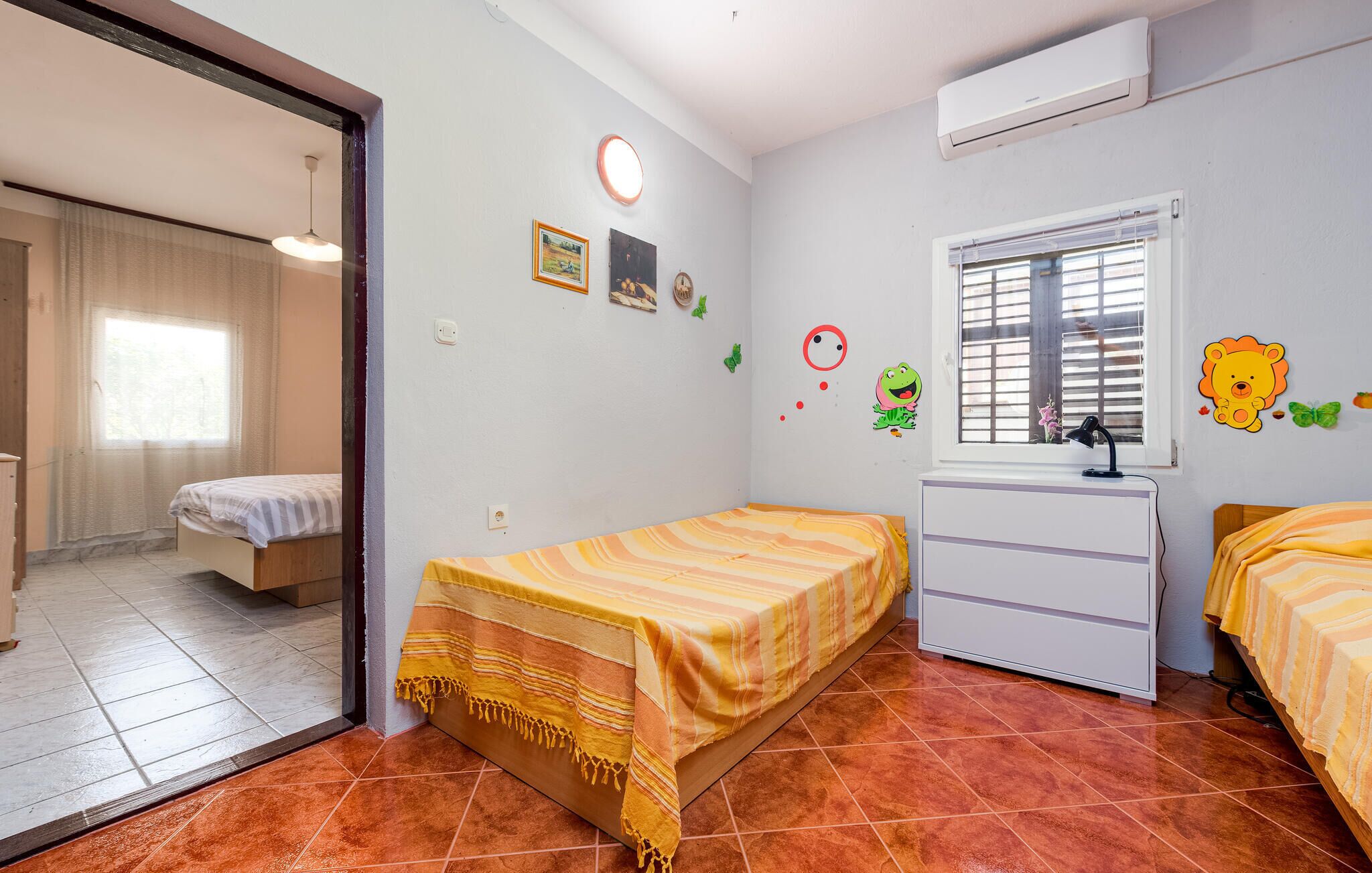 Une petite chambre bien éclairée, avec deux lits simples, un bureau avec une chaise et une fenêtre avec des stores.