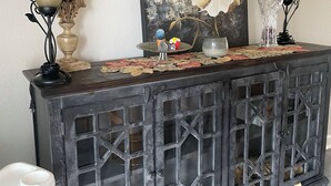 A dark wooden console table with a decorative top, flanked by two lamps with beige shades, a large floral painting, and a vase with a blue cactus and a small figurine. - Stillhouse Lake Fishing Retreat -Cedar Creek Ranch, 10 Acres + Boat Parking (Salado)