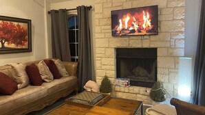 A cozy living room with a stone fireplace, a comfortable sofa, a coffee table, and a television displaying a fireplace scene. - Stillhouse Lake Fishing Retreat -Cedar Creek Ranch, 10 Acres + Boat Parking (Salado)
