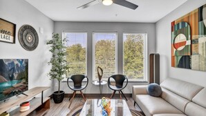 Ein modernes Wohnzimmer mit einer Couch, zwei Sesseln, einem Couchtisch und einem Fernseher. Es gibt drei Fenster mit Jalousien, einen Deckenventilator und ein Gemälde an der Wand.