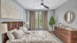 Ein Schlafzimmer mit einem Bett, einem Schminktisch, einem Deckenventilator, einem Fenster mit Vorhängen und einer Wand mit einer Dekoration.