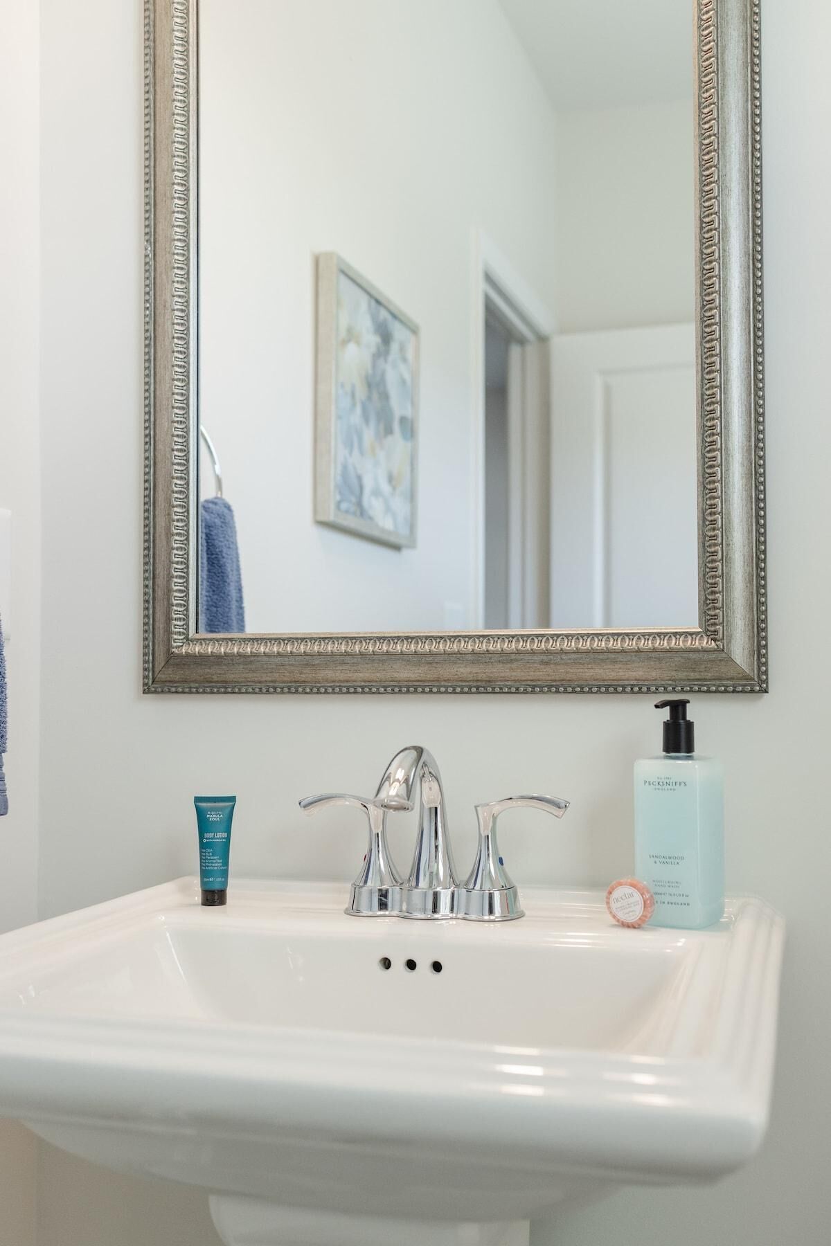 Un bagno con un lavandino bianco, un rubinetto argentato, uno specchio con cornice decorativa e articoli da toilette sul lavandino.