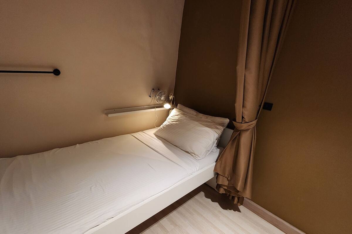 Ein kleines Zimmer mit einem Einzelbett, einem Nachttisch, einem Wandventilator und einem Vorhang.