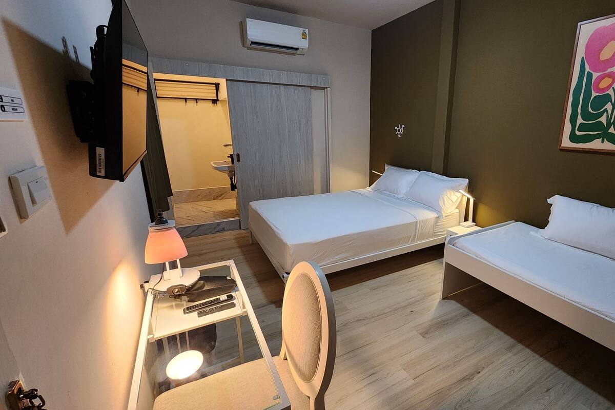 Ein Hotelzimmer mit zwei Betten, einem Schreibtisch mit Lampe, einem Sessel, einem Fernseher und einem durchgegangenen Tür angedeuteten Badezimmer.