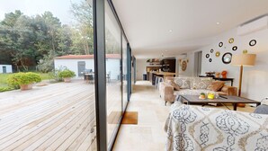 Amplia sala de estar con un gran ventanal corredizo que da a una terraza de madera, una cocina al fondo y un comedor con sofá y mesa de centro.