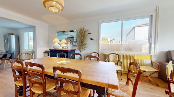 Een eetruimte met een grote houten tafel, zes stoelen, een lamp en uitzicht op buiten door een raam.