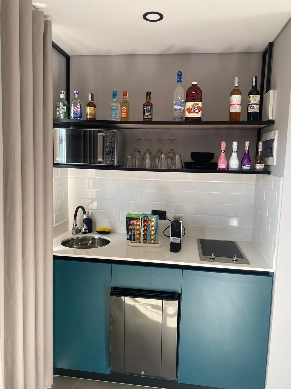 Cocina compacta con armarios color turquesa, un microondas, un fregadero y un refrigerador pequeño. Hay repisas arriba que contienen varias botellas y vasos.