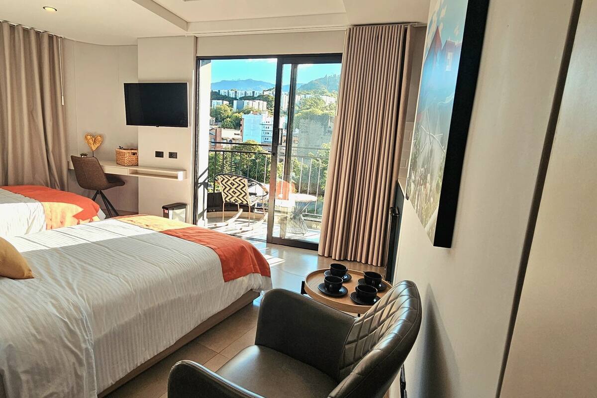 A modern hotel room with a large bed, a desk, a chair, and a balcony with a view of the city.