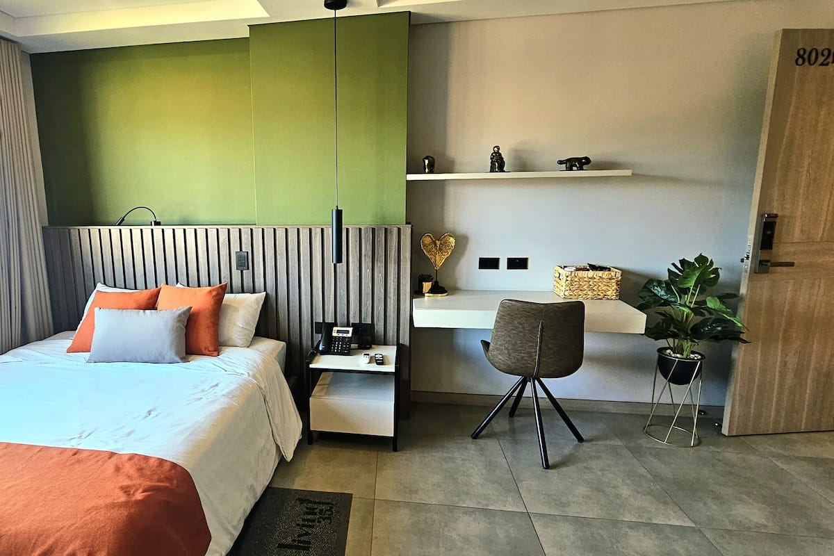A modern hotel room with a large bed, a desk, a chair, a potted plant, and a view of the city.