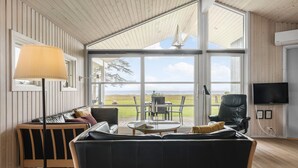 Uma sala de estar ampla com uma grande janela que oferece vista para o mar, um sofá confortável, uma mesa de jantar com cadeiras e uma televisão fixada na parede.