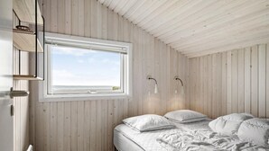 Quarto com teto de madeira, uma janela grande, cama com roupa de cama branca e duas luminárias fixadas na parede.