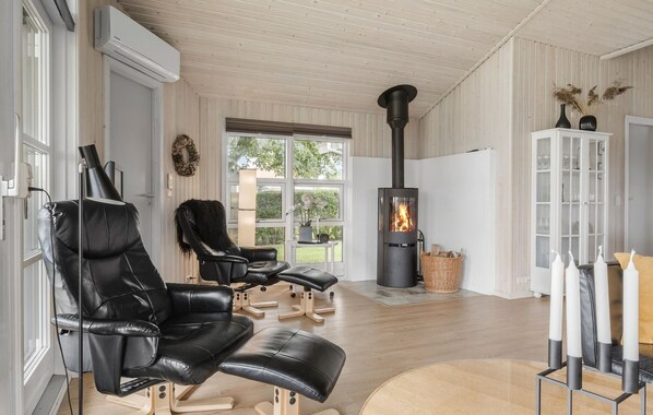 A modern living room with a wood-burning stove, leather chairs, a white cabinet, and a view through large windows.