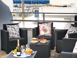 Un balcón con mobiliario de mimbre, una mesa con comida y bebidas, y vista a un puerto deportivo.