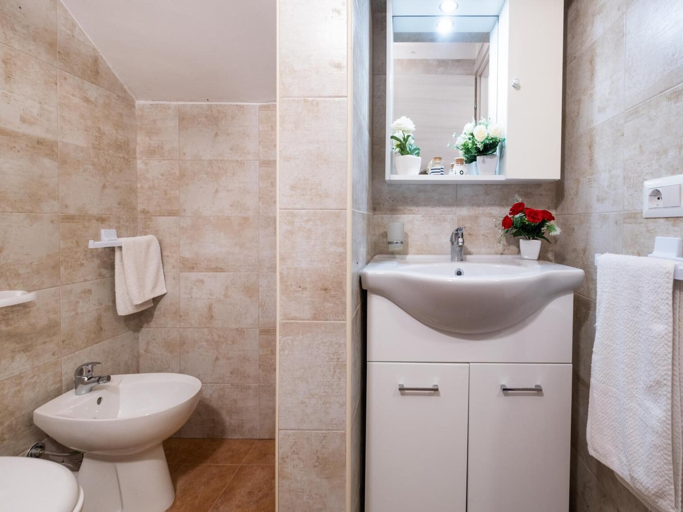 Une salle de bain équipée d’un lavabo, d’un bidet et de toilettes. Un miroir est placé au-dessus du lavabo, et une petite étagère y est fixée pour y déposer les articles de toilette.
