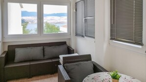 Ein sonnendurchfluteter Raum mit einem Korbsessel und einem kleinen Tisch, mit Blick auf einen See und Berge durch ein Fenster, sowie ein Fenster mit Jalousien.